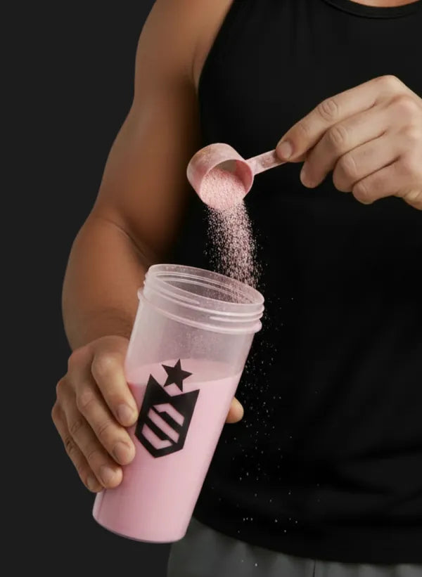 Mão despejando a medida de Whey Core sabor Morango numa coqueteleira com a bebida rosa