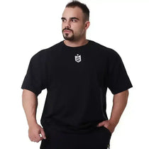 Homem musculoso vestindo camiseta preta oversized com estampa de escudo, fundo branco.