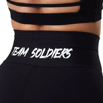 Detalhe de calça legging preta fitness cintura alta com escrita TEAM SOLDIERS na faixa