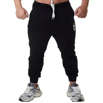 Calça jogger preta fitness masculina com cordão branco e tênis esportivo cinza sobre fundo branco.