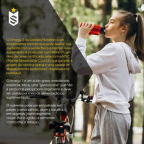 Jovem vestida de moletom cinza tomando suplemento energético com bicicleta ao fundo, ao ar livre.